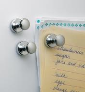 Three magnets on a fridge, one holding a shopping list and other papers