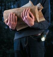 A person with a flashlight in a holster secured to their belt, carrying an armload of firewood