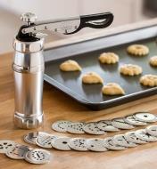 Aluminum Marcato Cookie Press sitting on a counter next to the included dies and a tray of cookies