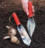 Spear-point trowel held in a hand and transplant trowel resting on soil next to garlic bulbs