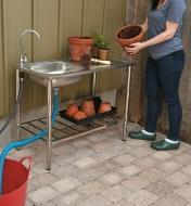 Femme tenant un pot près d'un évier de jardin en acier inoxydable dont le plateau et la tablette soutiennent des pots de différents formats