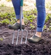 A woman wearing European garden clogs bears down on a garden fork to drive it into the soil 