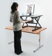 A woman stands as she works on a computer on a raised countertop desk lift on top of a table 