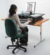A woman sits in a chair as she works on a computer on a lowered countertop desk lift on top of a table 