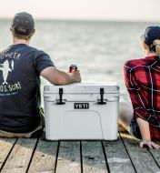 A man and woman sitting on a dock with a Yeti cooler between them