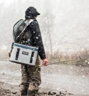 A man carrying a Yeti soft-sided cooler by the shoulder strap while fishing in a snowy location