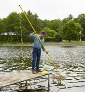 A man standing at the end of a dock rakes weeds from a pond