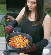 Woman wearing barbecue gloves holding cast-iron pan that she has removed from barbecue