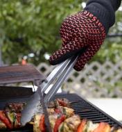 Wearing barbecue gloves while using tongs to turn food on barbecue.
