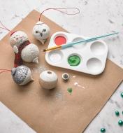 Decorated wooden ornaments lying on a table beside a tray of paint and a paintbrush