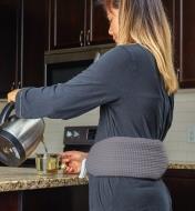 A woman wears the long hot-water bottle tied around her waist while standing in the kitchen making tea
