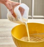 Personne pressant les bords d'un verre à mesure en silicone flexible pour ajuster l'écoulement de lait dans un bol à mélanger