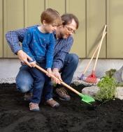 Adulte aidant un enfant qui creuse dans le jardin à l'aide de la bêche de l'ensemble d'outils de jardinage pour enfants