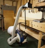 Rikon 1 hp portable dust extractor set up beside a workbench with the intake funnel close to a bench-top drill press