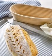 An oval bread is shown on a baking sheet next to an empty oval banneton