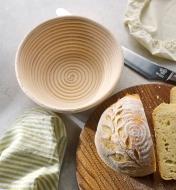 A round bread with a coil pattern and a scored wheat sheath design is shown on a wooden cutting board beside an empty round rattan banneton