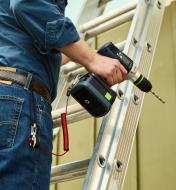 A person standing on a ladder with a cordless drill connected to their belt by a tool safety tether