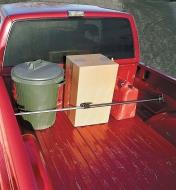 Barre de retenue à rochet posée dans la caisse d'une camionnette pour maintenir en place un bidon, une boîte en carton et une poubelle