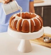 Pouring icing onto a Bundt cake sitting on a white cake stand