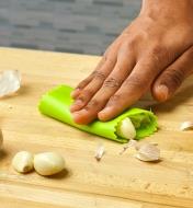 A hand presses on a green garlic peeler with garlic inside on a wooden cutting surface