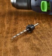 A drill bit with a stop collar sits in front of a drill on a table