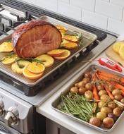 One baking pan with a rack and a ham on it and another baking pan filled with roasted vegetables
