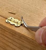 Mounting a small brass hinge on a wooden box, using forceps to hold a small nail while driving it