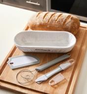 A freshly baked loaf of bread on a cutting board beside a proofing basket, whisk, scraper and lame