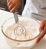 Personne mélangeant les ingrédients d'une pâte à pain dans un bol en verre avec un fouet danois