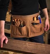 A woodworker inspects a piece of lumber while carrying various hand tools in a five-pocket waist apron
