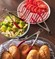 Tongs placed with rolls, a salad and a plate of meat and tomatoes