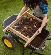 A sifter filled with earth is placed over a wheelbarrow