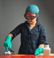 A woman wearing chemical splash goggles while using chemicals to strip the finish from wood
