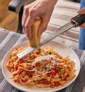 Using a handled stainless-steel rasp to grate parmesan cheese onto a plate of pasta