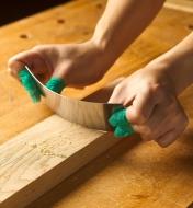 A person using a wood scraper on a workpiece has their fingers wrapped in high-friction guard tape 