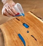 A woodworker pours blue epoxy from a silicone mini-measure to fill a recess in a live-edge board