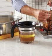 Tasse à dégraisser contenant du jus de cuisson sur un comptoir à côté d’une dinde rôtie sur une lèchefriteer