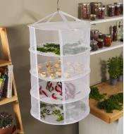 A four-tier herb dryer hanging in a kitchen with herbs and vegetables spread out to dry