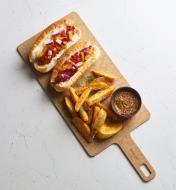 A cut & serve board with hotdogs, wedge potatoes and a small dish on it