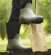 A person wearing garden boots using their foot to push a spade into the ground