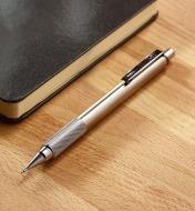 The stainless-steel mechanical pencil sits on a wood surface next to a notebook