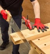 A man wearing Flex Grip gloves uses a handsaw to cut a wooden board clamped to a workbench