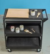 A brush and an open can of finish beside a wooden cutting board placed on a workshop tool cart