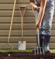 A child uses the digging fork to move soil, while the rake and spade are behind, leaning up against siding