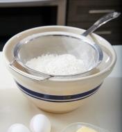 A sifter with flour in it resting on a mixing bowl