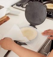 Personne soulevant une feuille de papier parchemin qui recouvre une tortilla sur la plaque inférieure d'une presse à tortilla