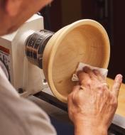 A woodturner uses a paper towel to apply Yorkshire Grit abrasive paste onto a wooden bowl on a lathe