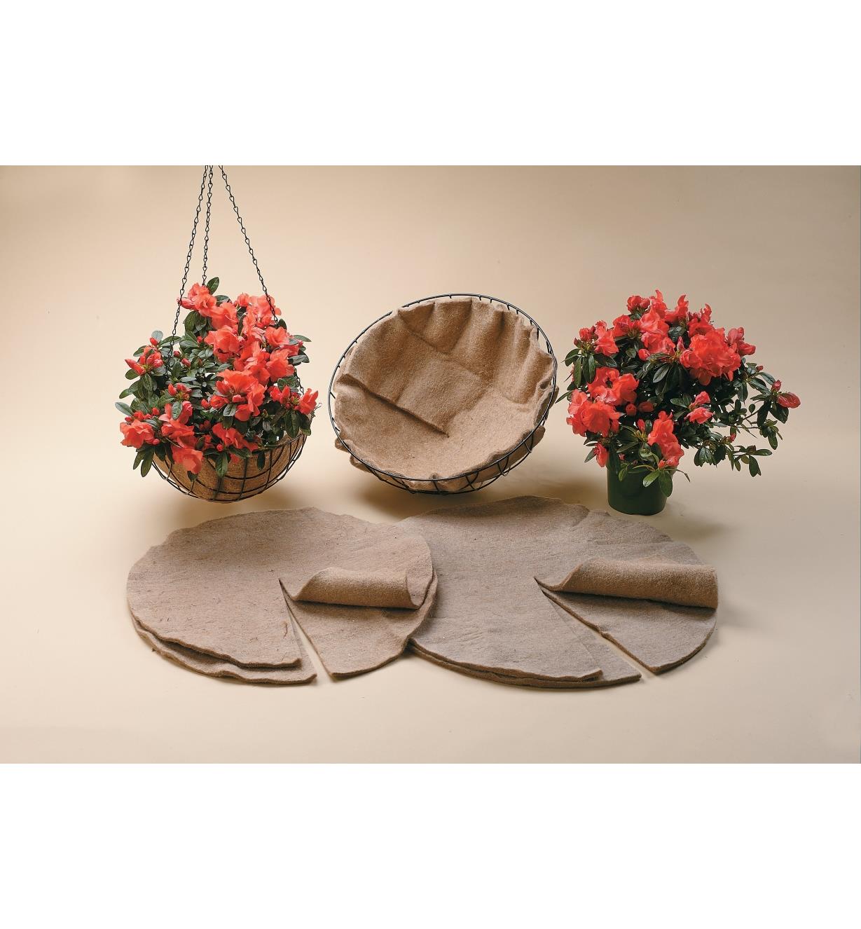 Both sizes of basket liners sitting in front of two baskets with liners in them, one filled with a flowering plant and hanging from chains, alongside a flowering potted plant