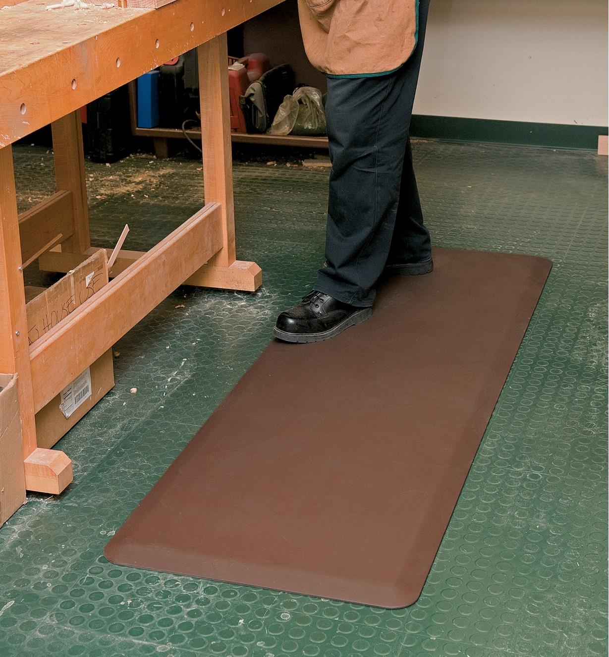 A person stands on a 24" x 72" brown Stationary Mat beside a workbench