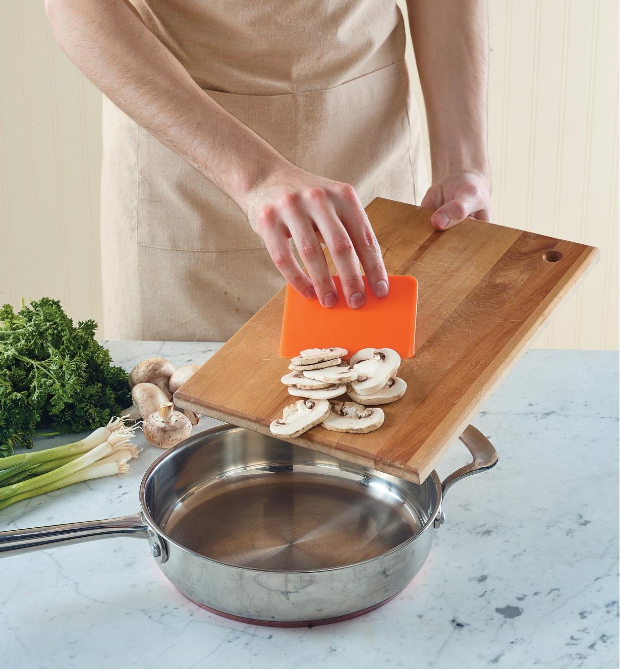 Using the food prep scraper to transfer sliced mushrooms from a cutting board into a saucepan
